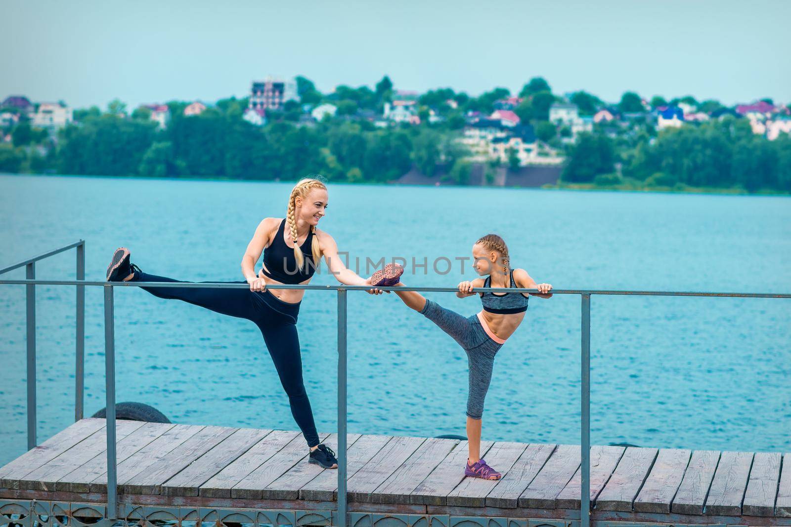 Woman and child are doing sport exercises stretching their legs on the pier the lake