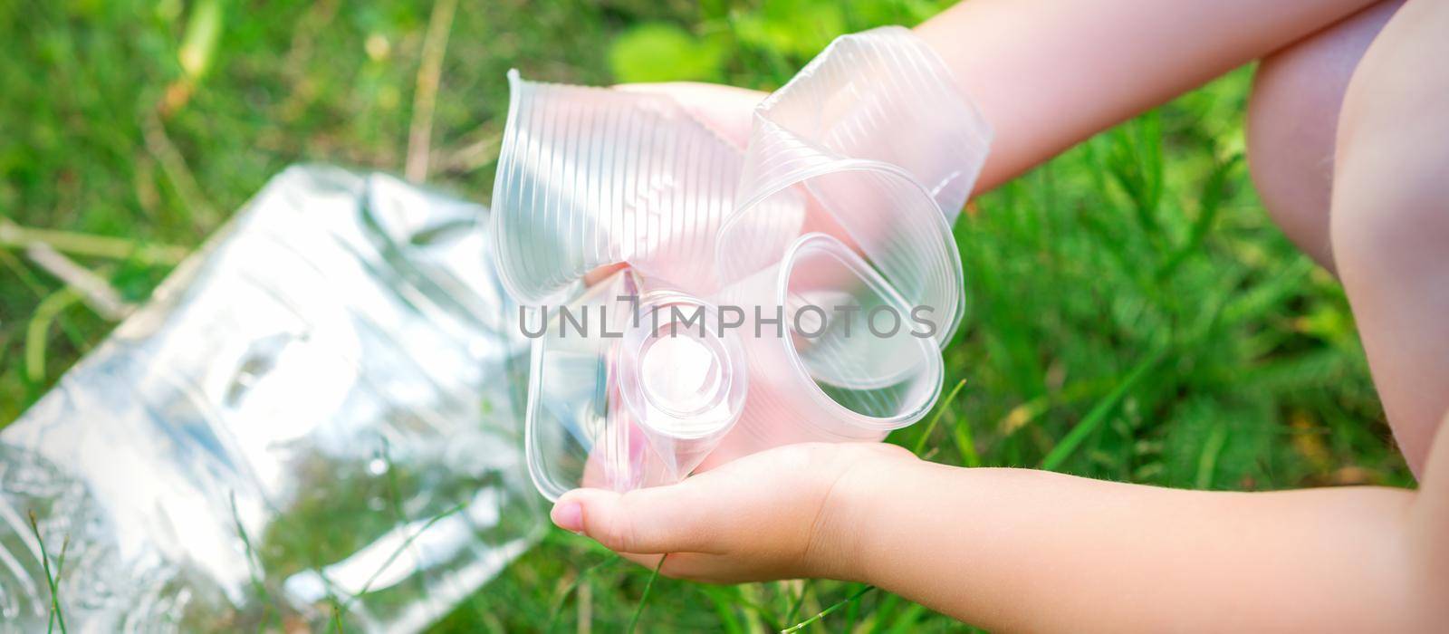 Child's hands clean the park from used plastic utensils in the grass