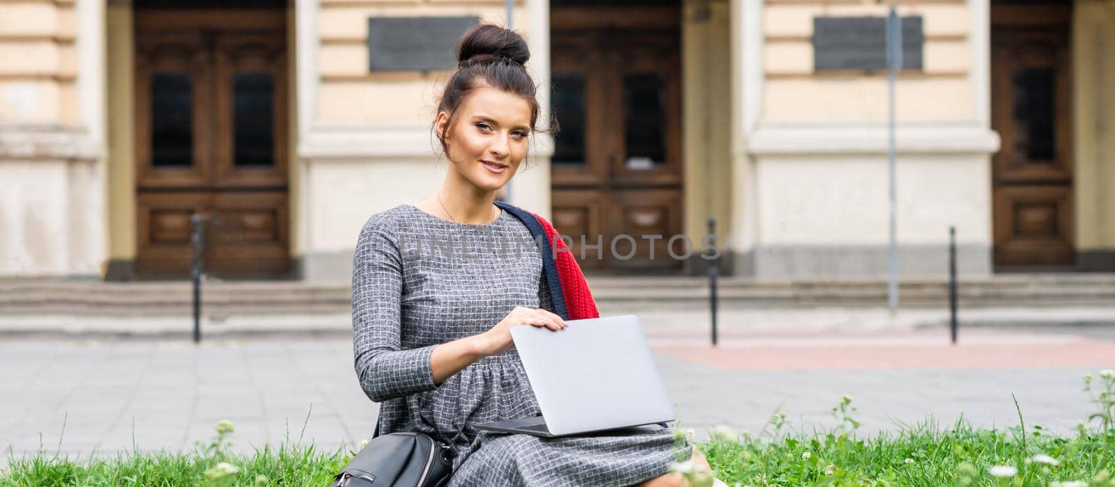 Portrait of a beautiful young smiling student studying with a laptop on the grass near the university building