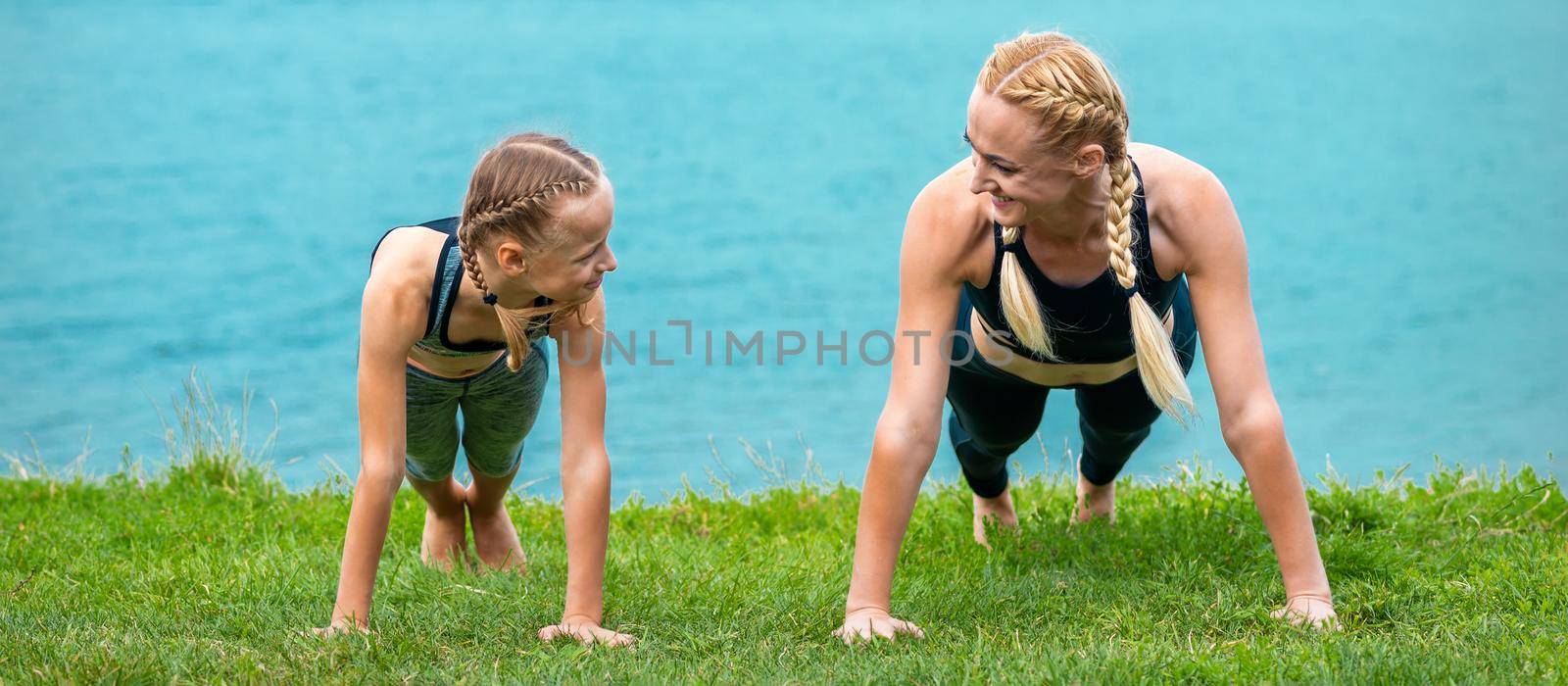 Woman and girl doing push-up exercise on the grass near lake outdoors
