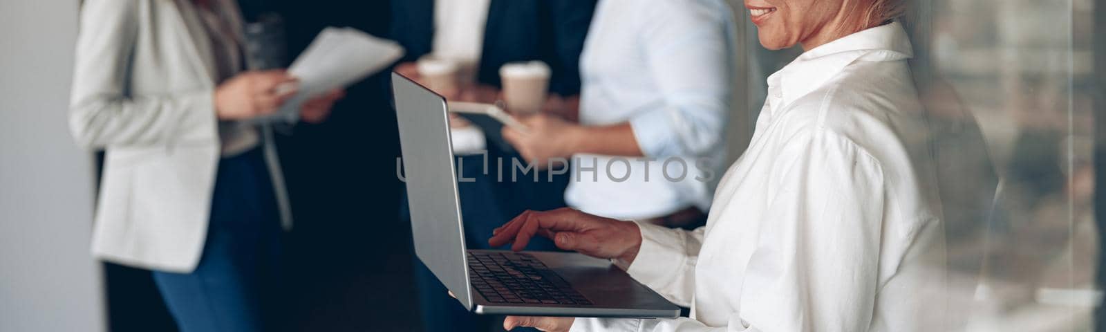 mature confident businesswoman at office with group of colleagues on background, working on laptop by Yaroslav_astakhov