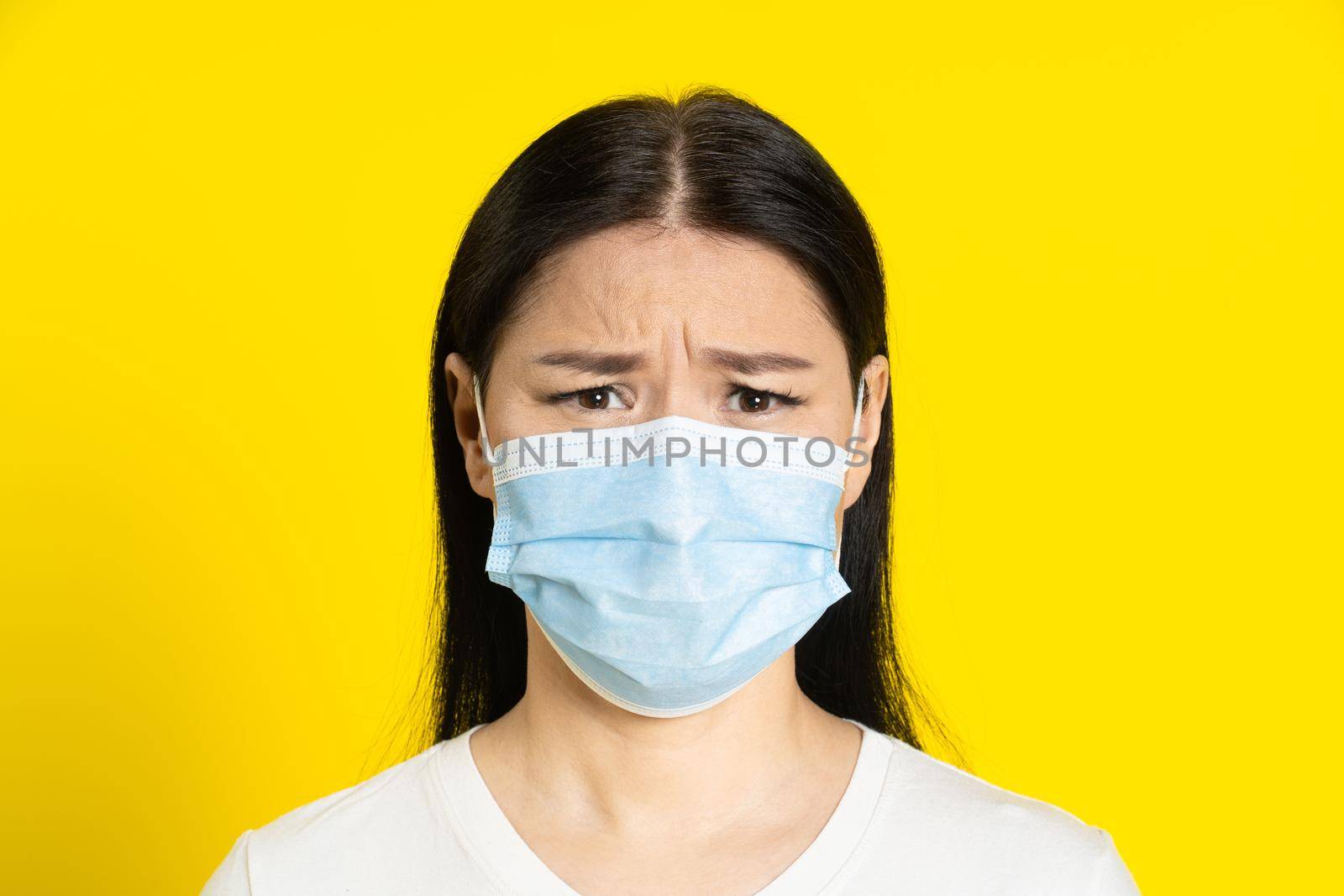 Angry, sad of pandemic mature asian woman wearing medical face mask coronavirus or monkeypox prevention. Charming middle age woman in white t-shirt and medical mask on yellow background.