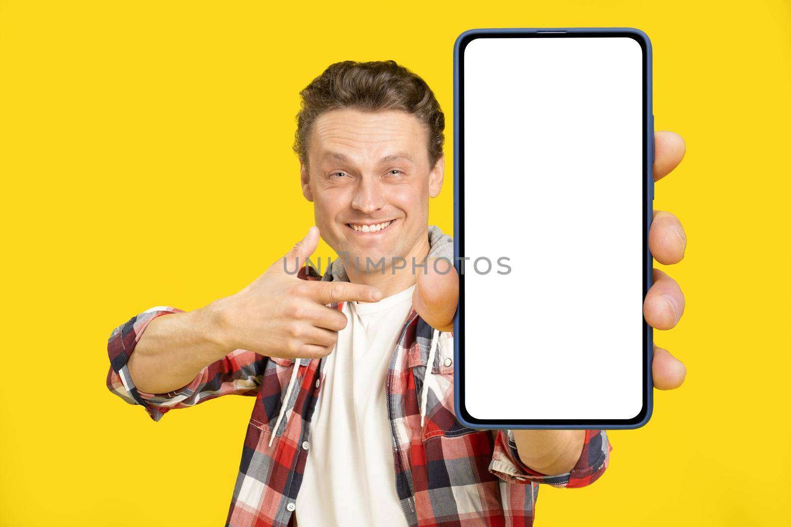 Pointing at giant, huge smartphone with white empty screen blond man, wearing red plaid shirt. Man with phone display mock up isolated on yellow background. Mobile app advertisement.