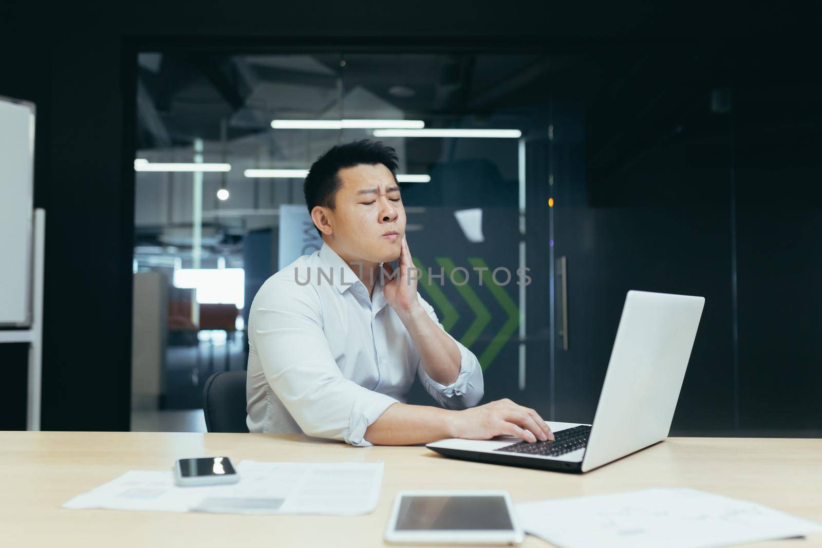 Asian man working in modern office, businessman having severe toothache, sick employee at work by voronaman