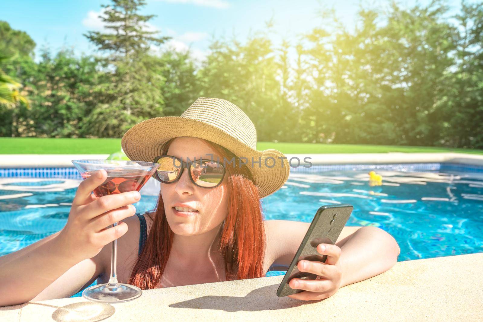 young woman relaxing inside the pool, looking at her mobile phone. girl on holiday sharing the moments on her social networks. concept of summer and free time. outdoor garden, natural sunlight.