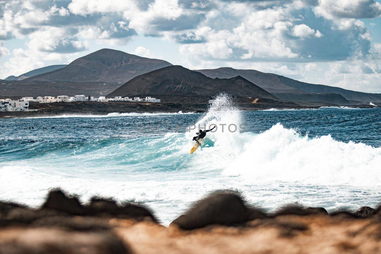 Wild rocky coastline of surf spot La Santa Lanzarote, Canary Islands, Spain. Surfer riding a big wave in rocky bay, volcano mountain in background