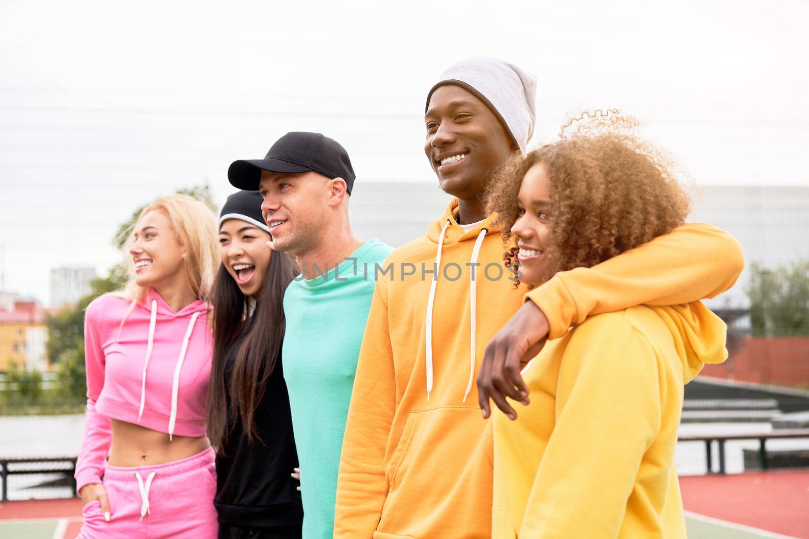 Group of multiethnic friends in colorful sportsuits walking together outdoor at sport ground in city park, looking aside and laughing