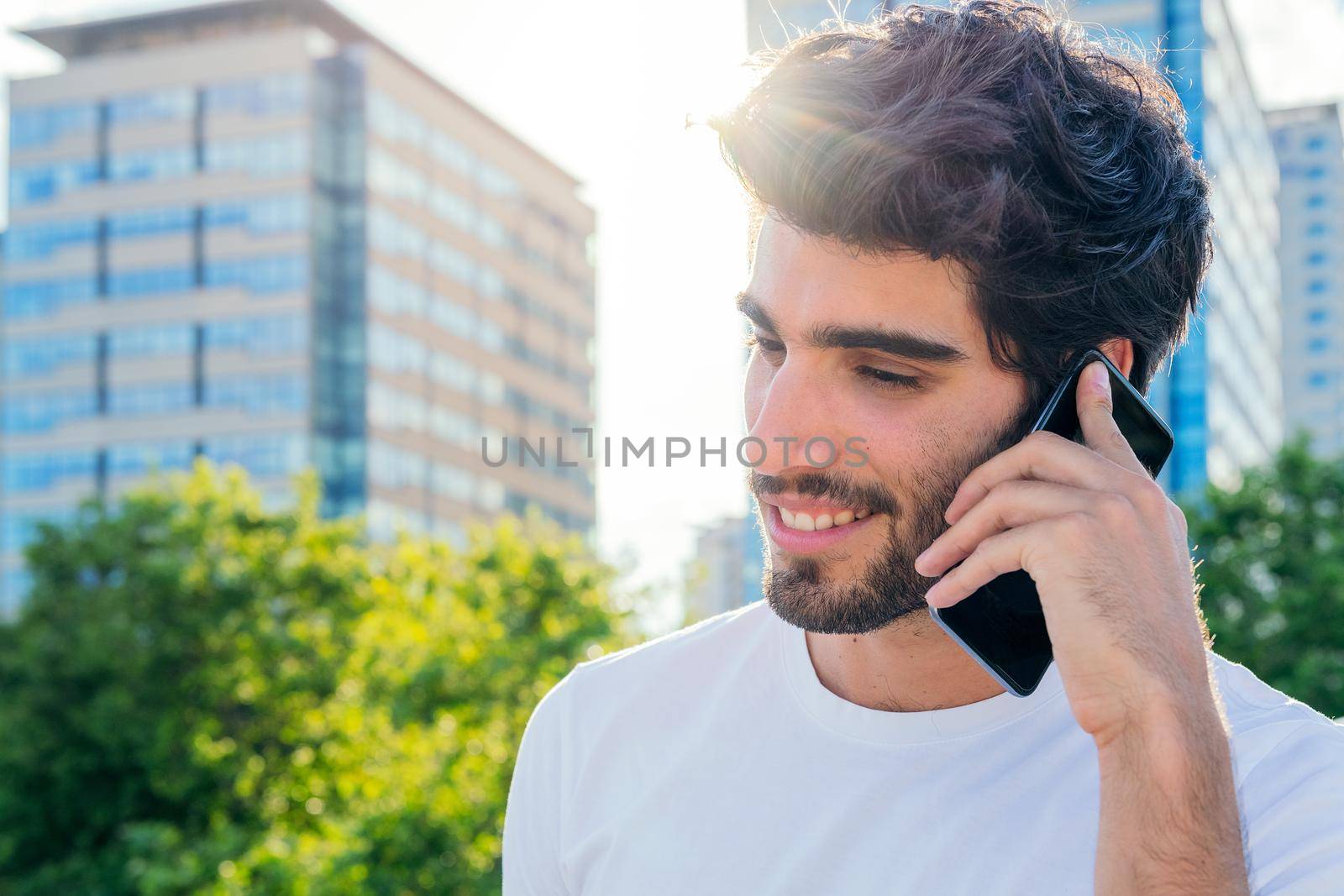 portrait of a handsome young man smiling and talking on his phone at sunset in front of the city buildings, concept of technology and urban lifestyle, copy space for text