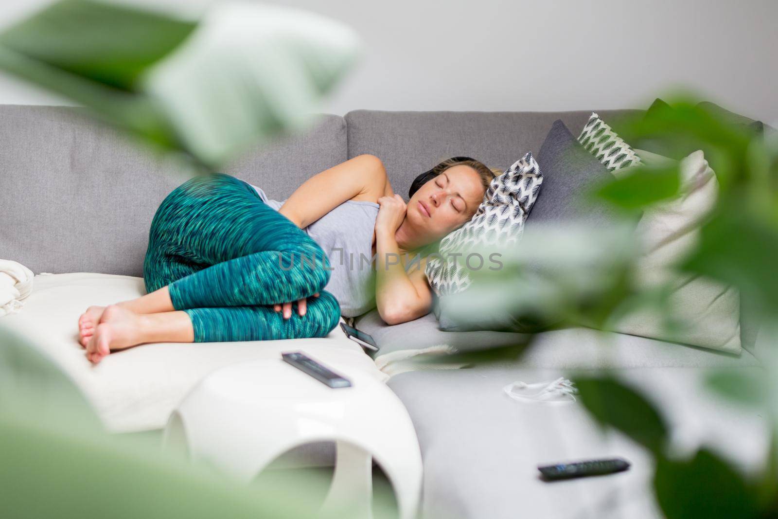 Young casual woman lying on couch cushion with eyes closed, relaxing on cozy sofa pillow, relaxed girl taking nap at home, wearing headphones, listening to music or podcast. by kasto