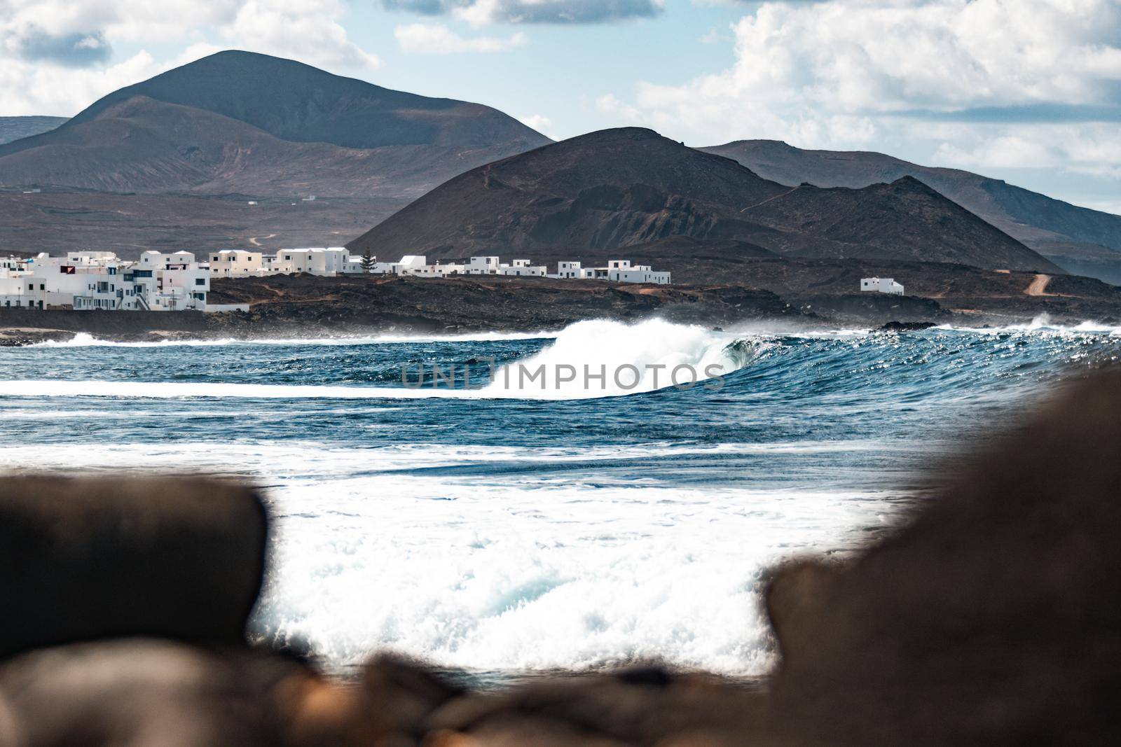 Wild rocky coastline of surf spot La Santa Lanzarote, Canary Islands, Spain. Surfer riding a big wave in rocky bay, volcano mountain in background. by kasto