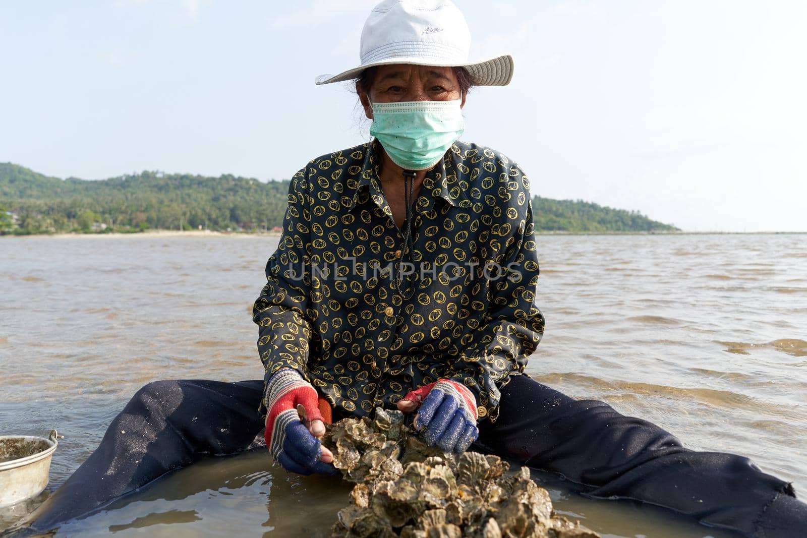 Ko Phangan, Thailand, March 15, 2022: old woman with protective mask searching for clams on the sea