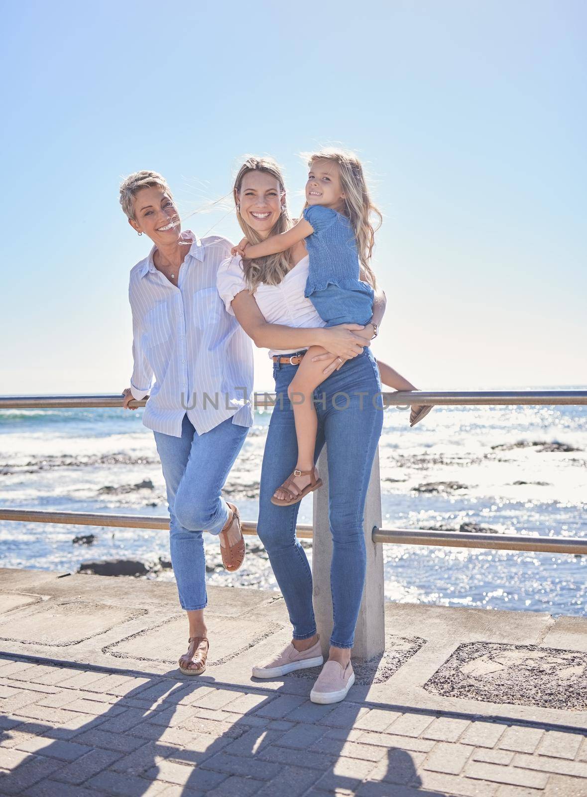 Full length of female family members posing together at the beach on a sunny day. Grandmother, mother and granddaughter standing together on seaside promenade. Multi-generation family of women and little girl spending time together.