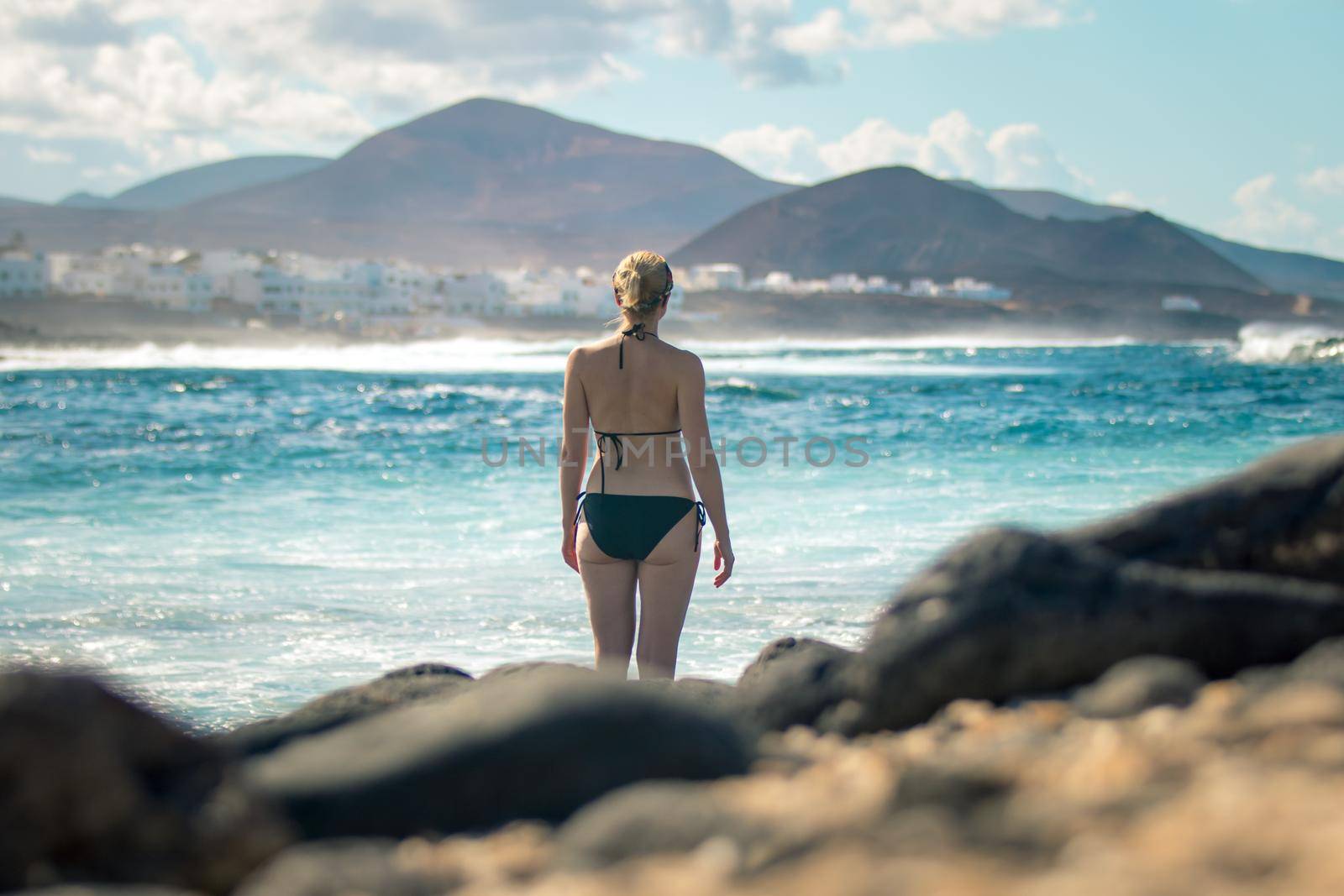 Female tourist at wild rocky beach and coastline of surf spot La Santa Lanzarote, Canary Islands, Spain. La Santa village and volcano mountain in background. by kasto