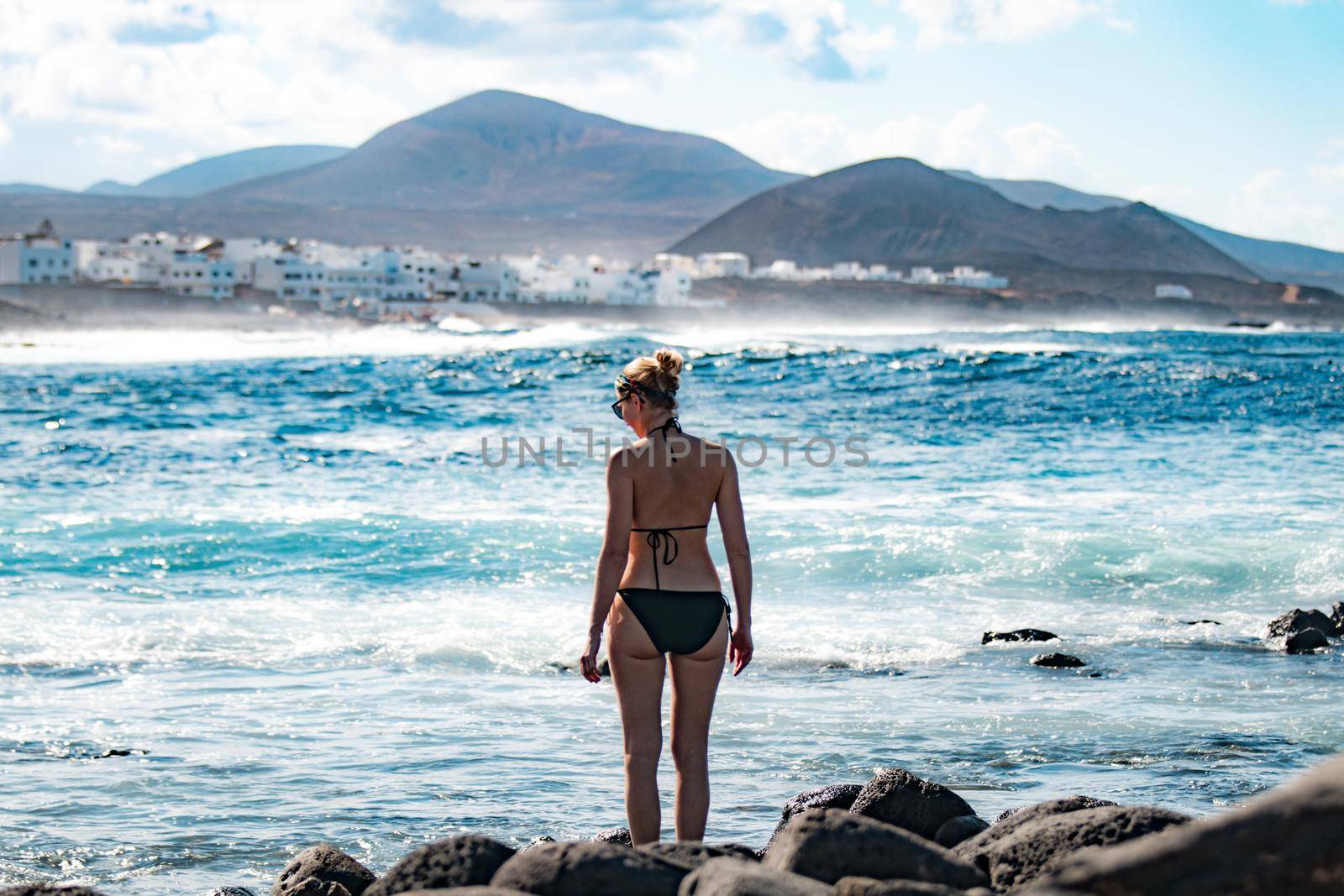 Female tourist at wild rocky beach and coastline of surf spot La Santa Lanzarote, Canary Islands, Spain. La Santa village and volcano mountain in background. by kasto