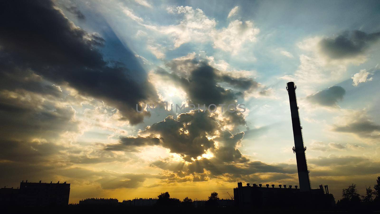 The chimney of the boiler room and the silhouettes of buildings against the backdrop of beautiful clouds and the sun behind them. Sunlight penetrates through the clouds by ProjectStockman