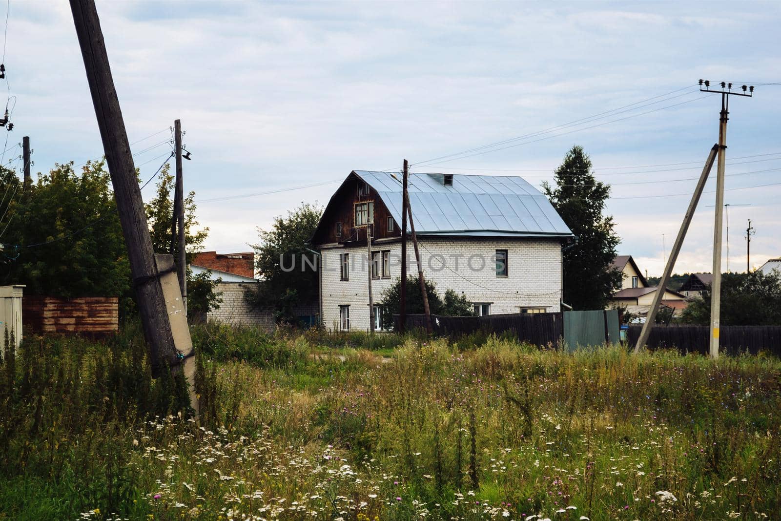 Glade with grass and flowers and an old two-story house in a Russian village under a cloudy sky by ProjectStockman