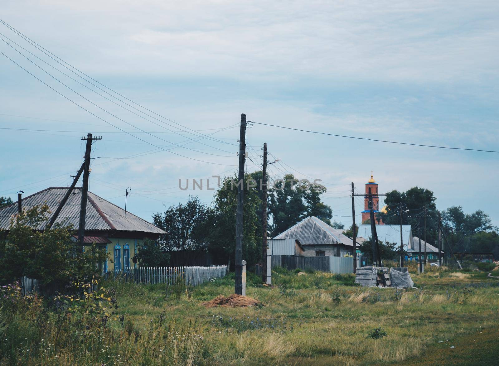 Old houses and power line poles against the backdrop of an Orthodox church under a blue cloudy sky in a village in Russia by ProjectStockman