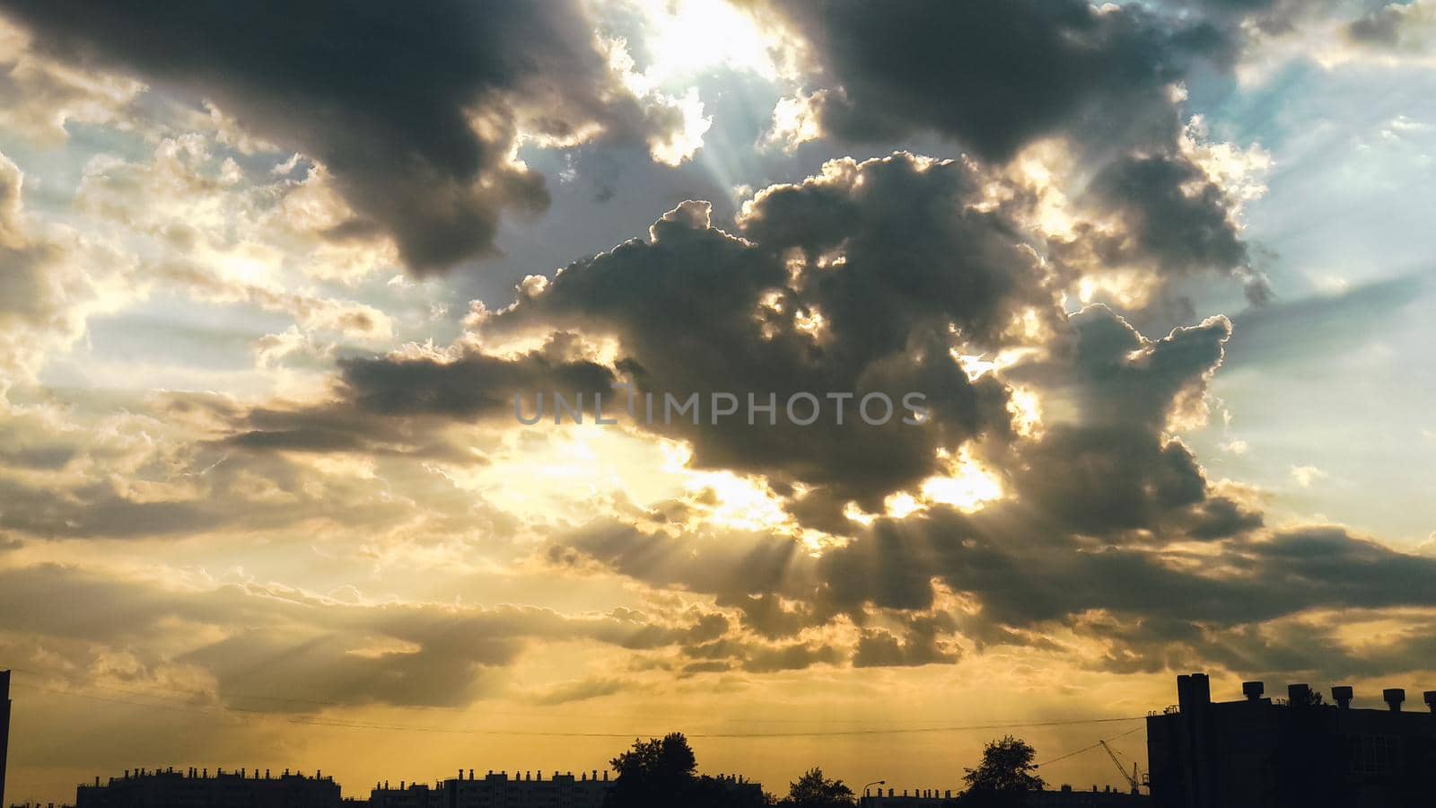 The sun in the clouds casts the sun's rays across the beautiful sky above the silhouettes of buildings and trees by ProjectStockman