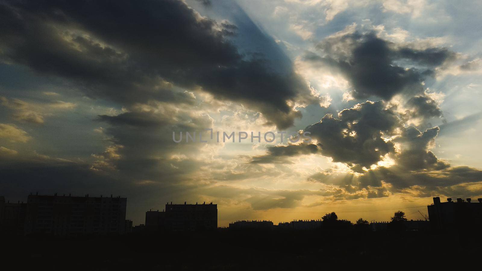 The sun behind the cloud casts the sun's rays across the beautiful sky above the silhouettes of buildings by ProjectStockman