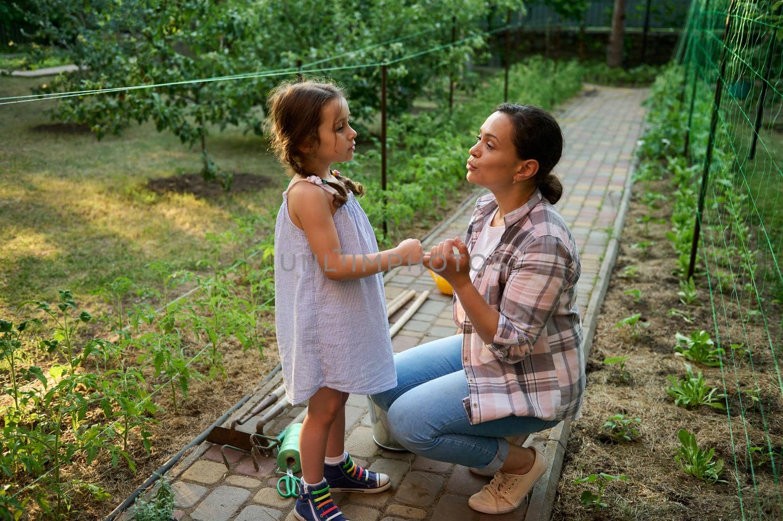 Multiethnic woman, loving mother and her lovely little daughter enjoying gardening at sunset time in the eco farm, planting and caring for seedlings. Install love and care for nature since childhood