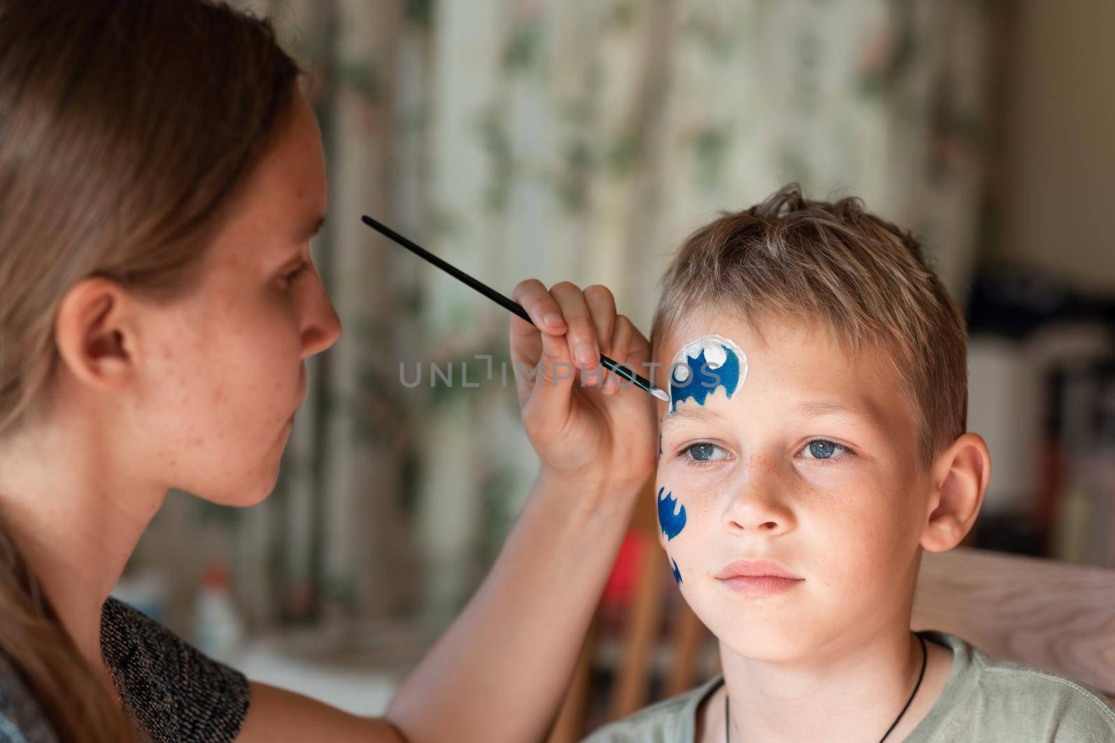 Child animator, artist's hand draws face painting to little boy. Child with funny face painting. Painter makes blue batman mask on boy's face. Face painting at children holiday, event, birthday party, entertainment.