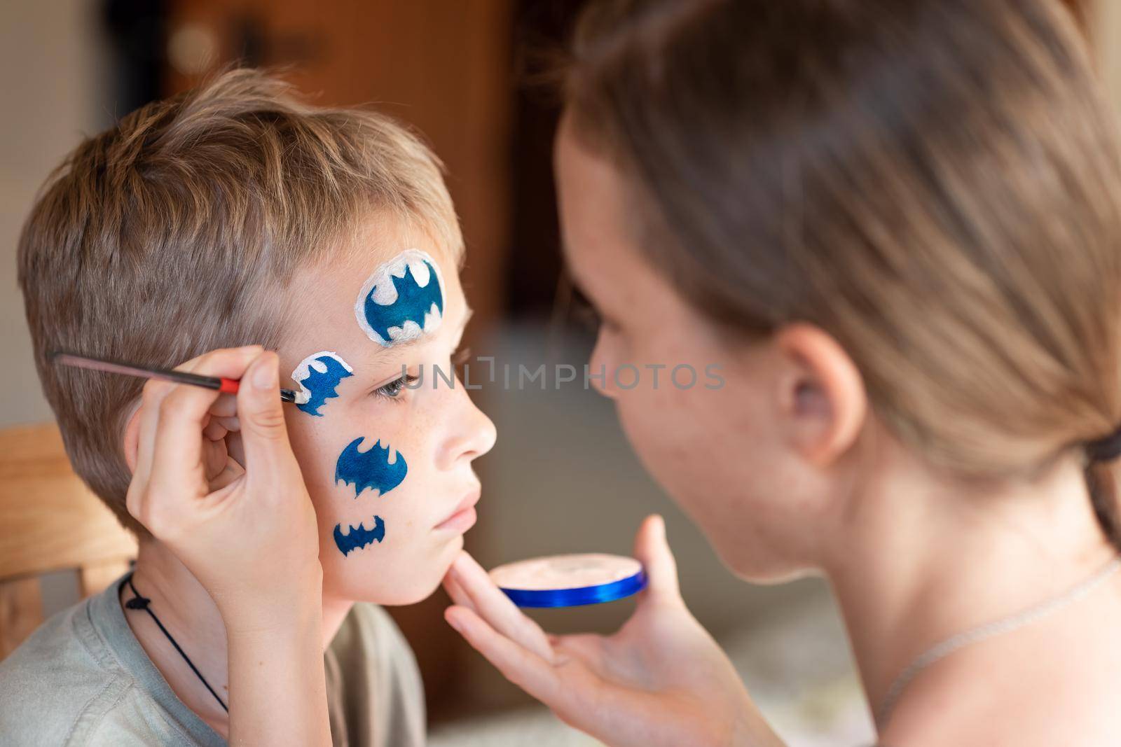 Child animator, artist's hand draws face painting to little boy. Child with funny face painting. Painter makes blue batman mask on boy's face. Face painting at children holiday, event, birthday party, entertainment.