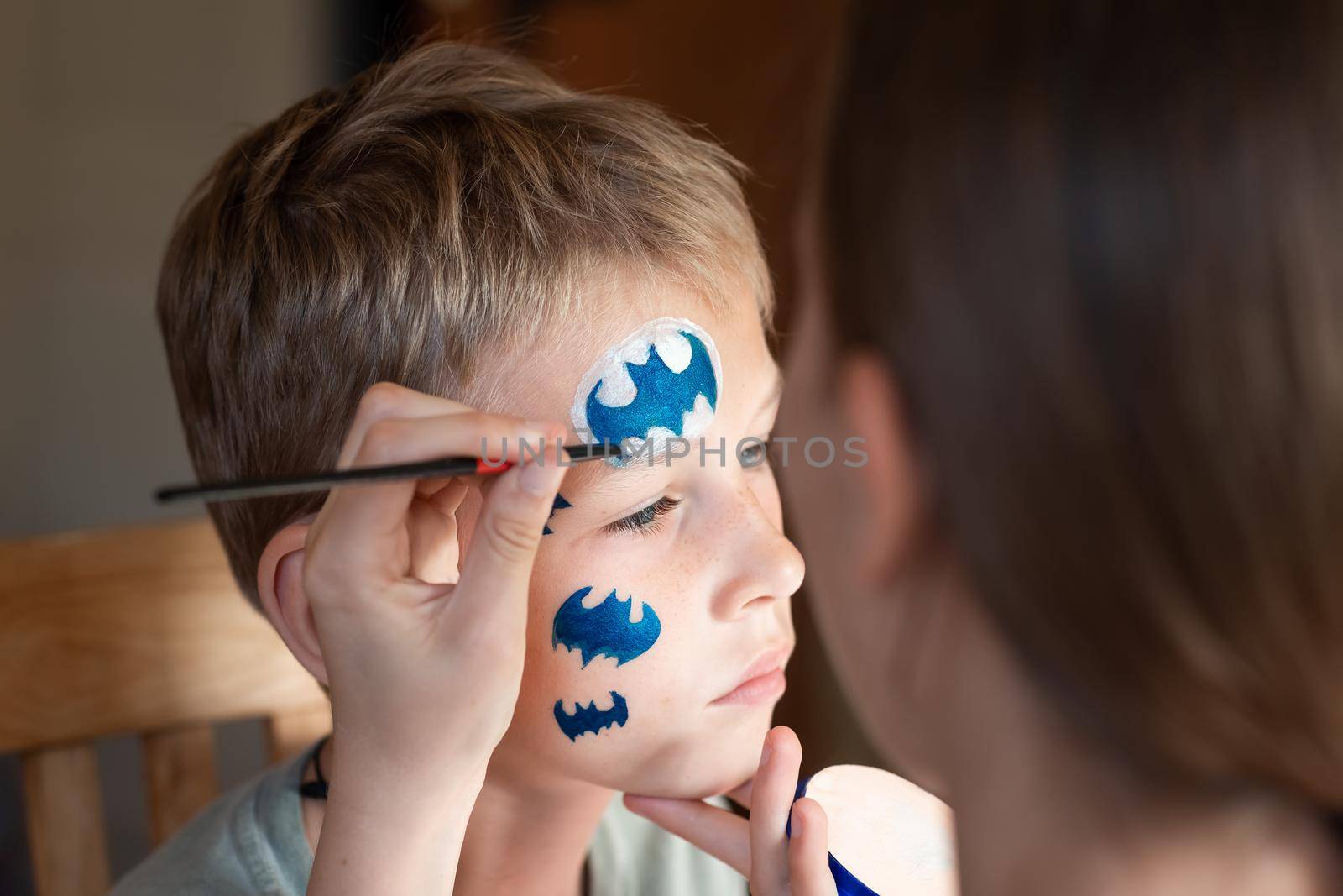 Child animator, artist's hand draws face painting to little boy. Child with funny face painting. Painter makes blue batman mask on boy's face. Face painting at children holiday, event, birthday party, entertainment.