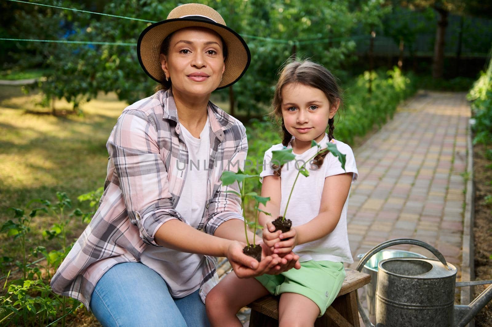 Pleasant young woman, mom and her daughter holding soil with growing cucumber seedlings in their hands, looking at camera. Family agribuisness, eco farming. Concept of World Environment Day. Ecology.