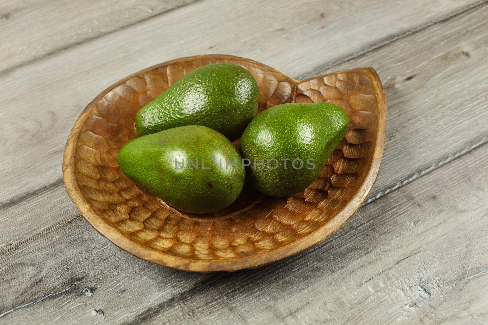 Three whole green avocados on wooden carved bowl.