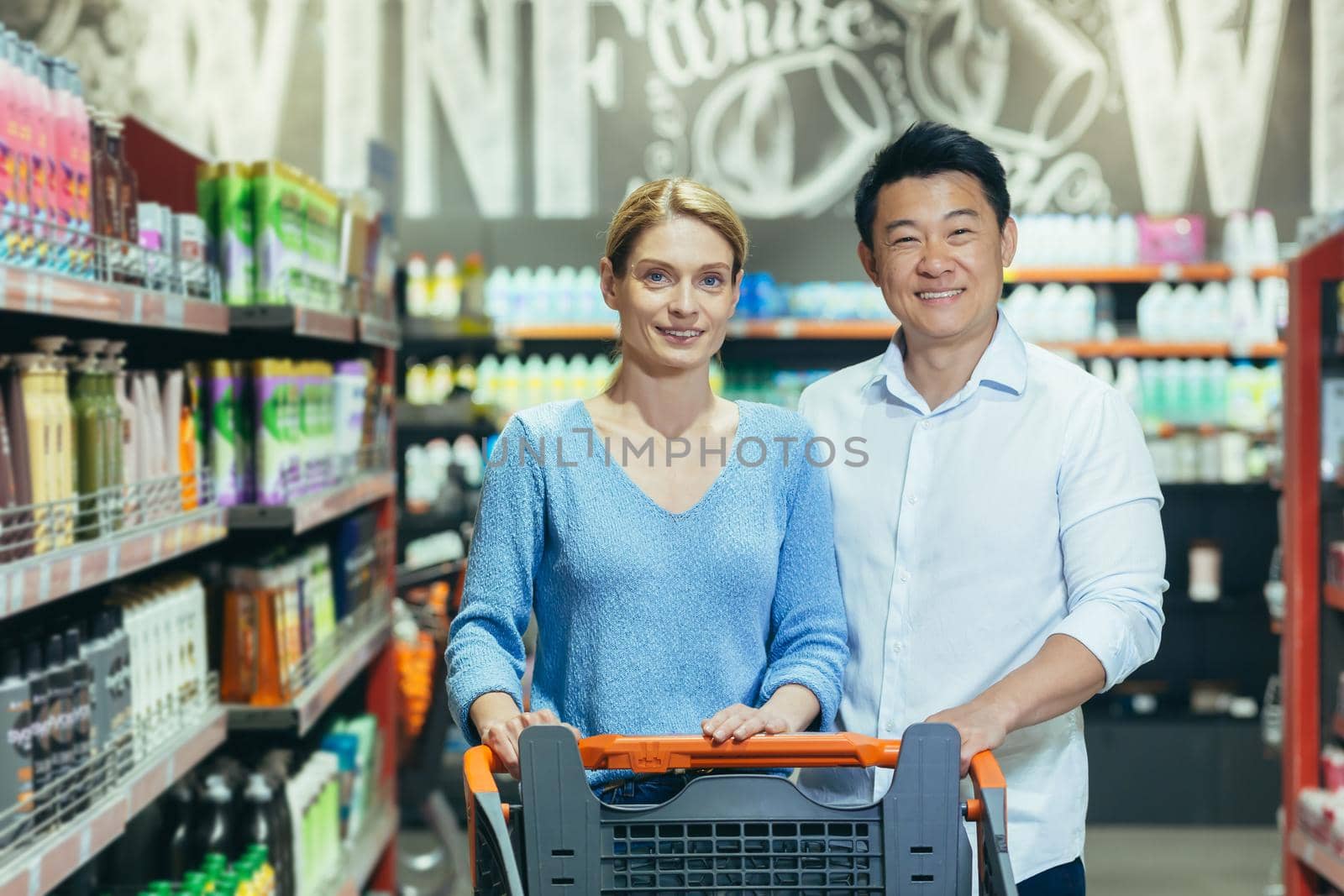 Portrait of happy shoppers in supermarket, interracial couple Asian man and woman, smiling and looking at camera, among shelves with goods