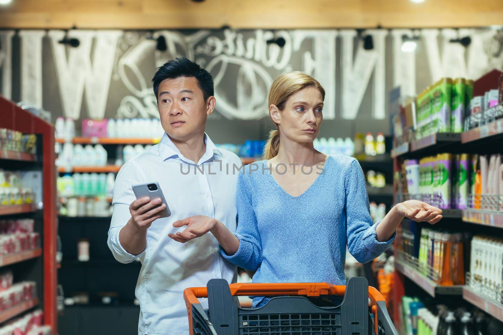 A couple of buyers, a man and a woman, confused in the supermarket, cannot choose the necessary product, an Asian man with a phone