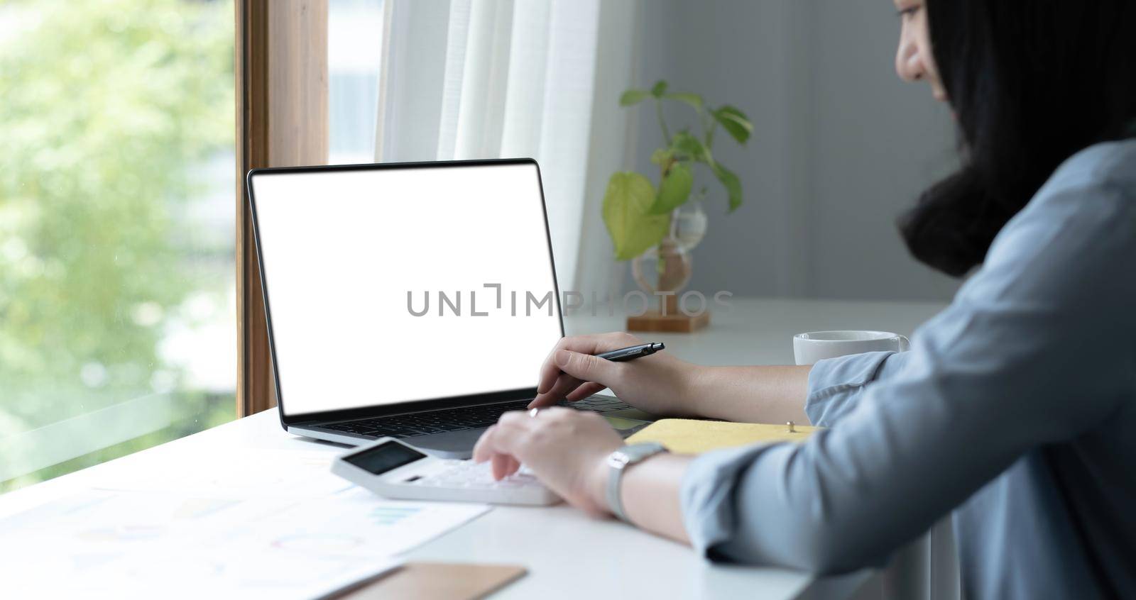 Mockup image of a woman using laptop with blank white screen on wooden table in office.
