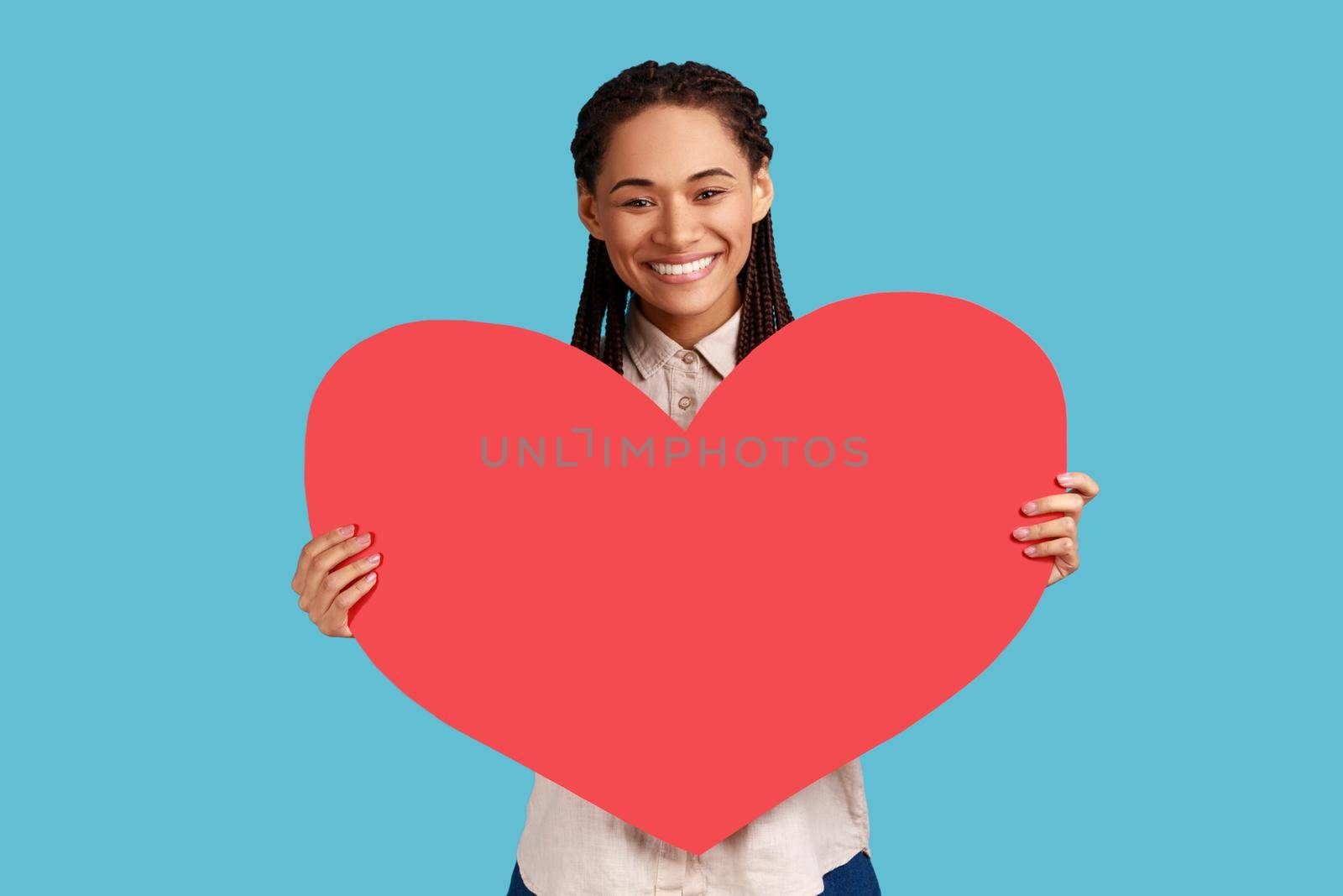 Portrait of attractive smiling woman with black dreadlocks holding big red heart, expressing positive romantic emotions, wearing white shirt. Indoor studio shot isolated on blue background.