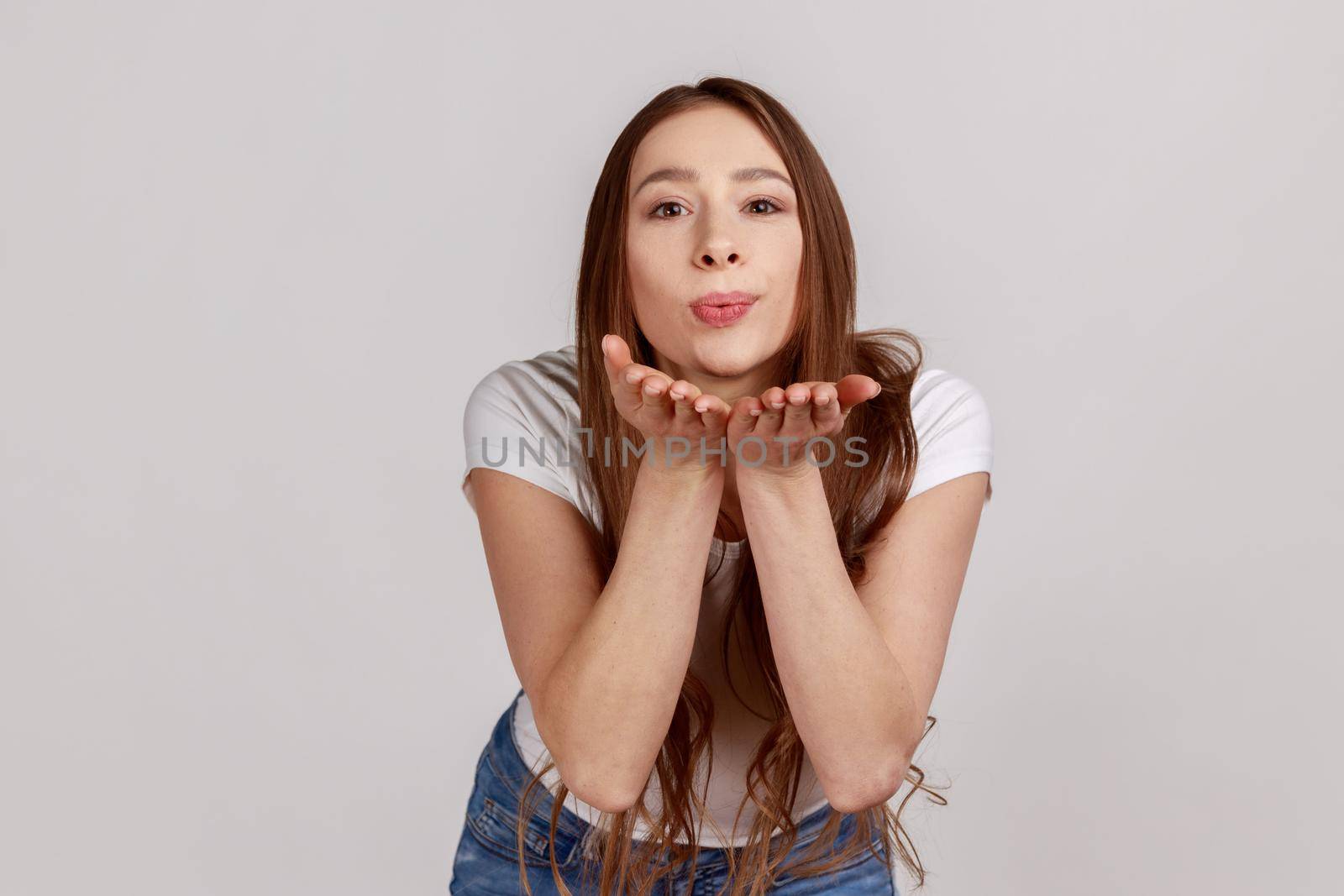 Catch my kiss. Lovely romantic woman looking at camera, sending air kissing over palms hands, expressing love, wearing white T-shirt. Indoor studio shot isolated on gray background.