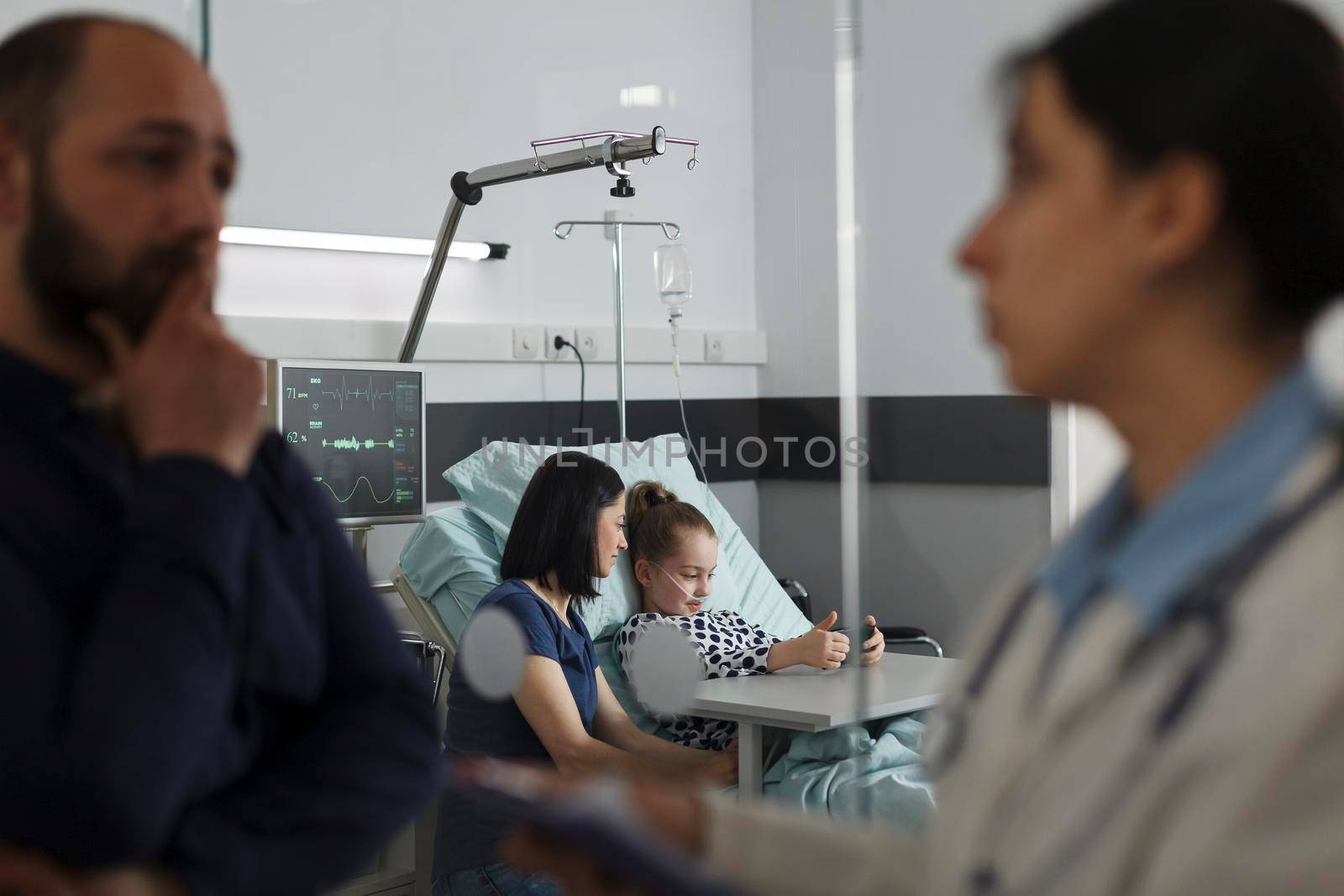 Sick girl playing games on smartphone with mother while father talking with expert pediatrist by DCStudio