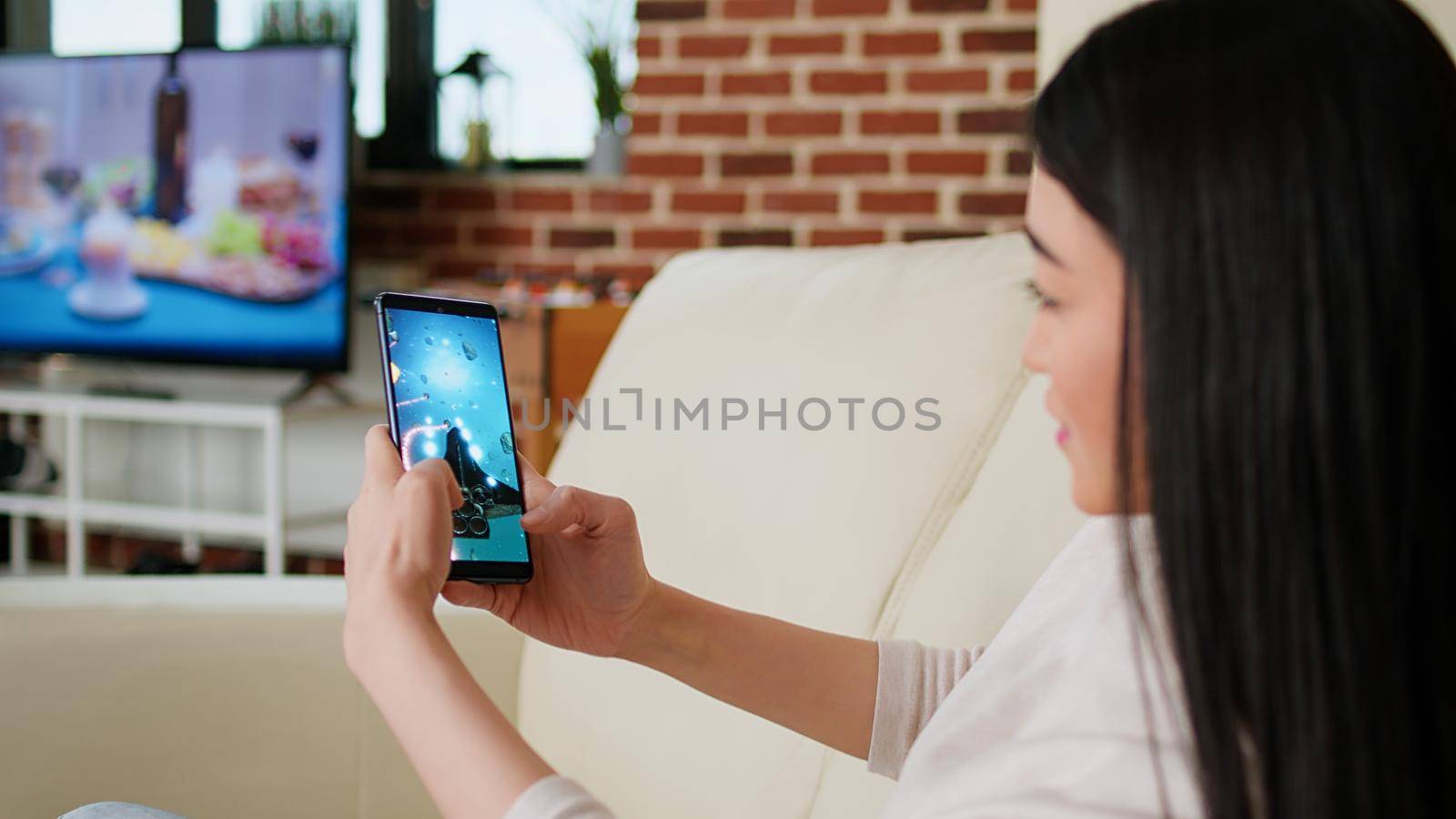Playful asian woman playing space shooter game on smartphone device while working remotely from home. Childish young adult person enjoying mobile gaming while sitting on sofa inside apartment.