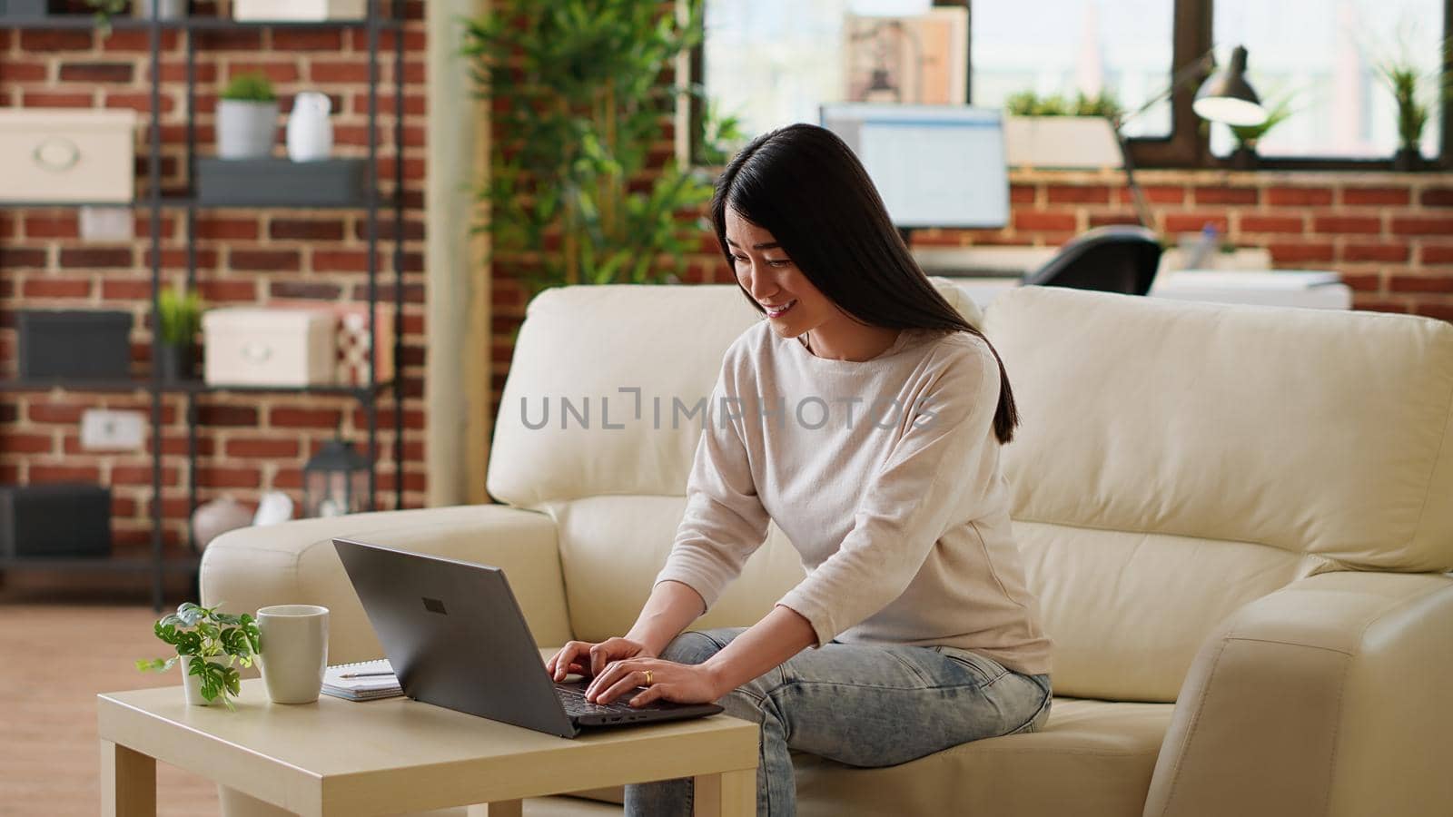 Young adult person doing remote work on modern portable computer while at home. Smiling heartily woman at home working remotely on laptop while sitting on sofa in living room.