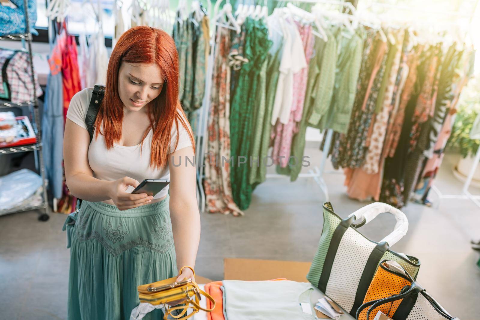 young smiling redhead girl shopping in a small clothing shop, taking pictures with her smart phone of the clothes, for her social networks. girl spending time shopping. concept of shopping.. natural light from the window of the window.