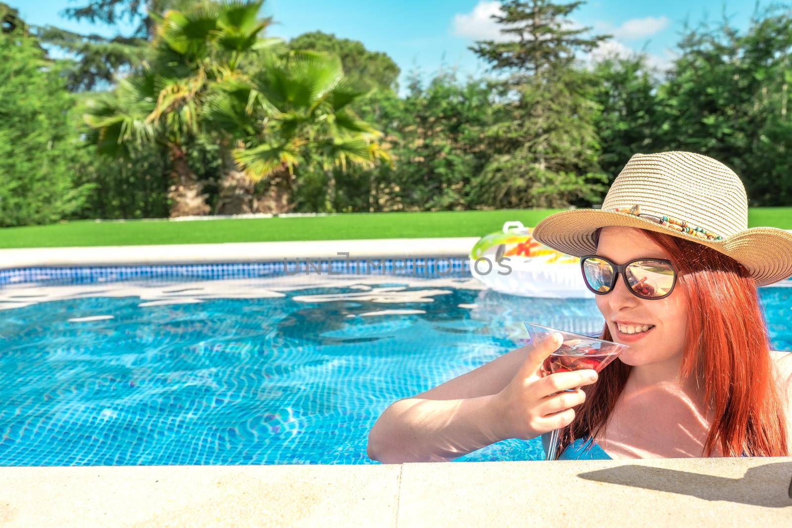 young woman with hat and sunglasses, having a refreshing drink inside the swimming pool. young girl on summer holiday sunbathing by the pool. concept of summer and free time. outdoor garden, natural sunlight.