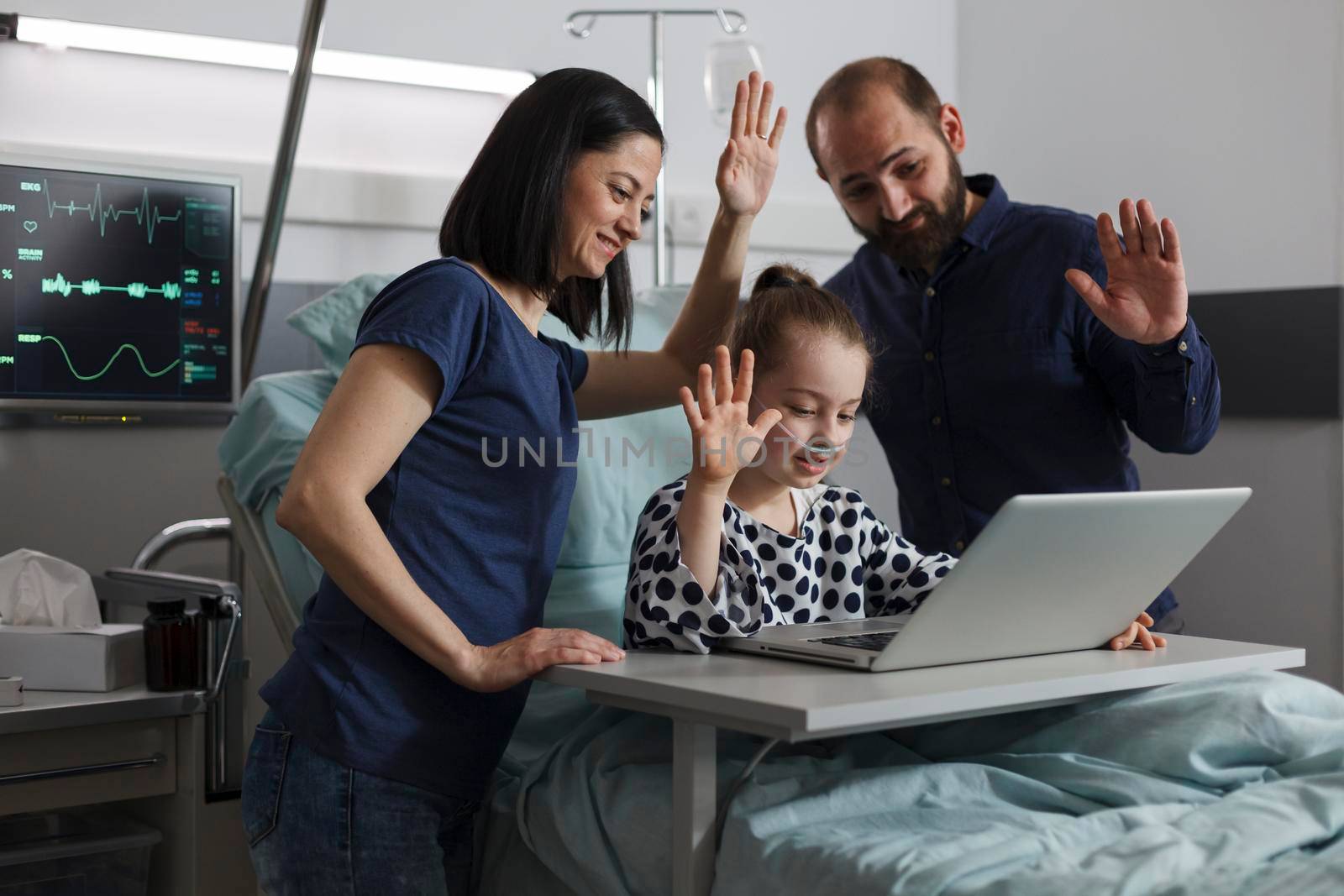 Ill little girl under treatment talking with relatives on online call while sitting with loving parents inside hospital ward. Mother and father standing beside daughter while waving at videocall.