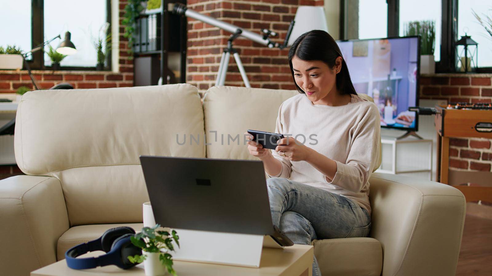 Playful woman sitting on sofa at home while playing arcade games on smartphone. Competitive looking young adult person enjoying mobile gaming while relaxing in living room on couch.