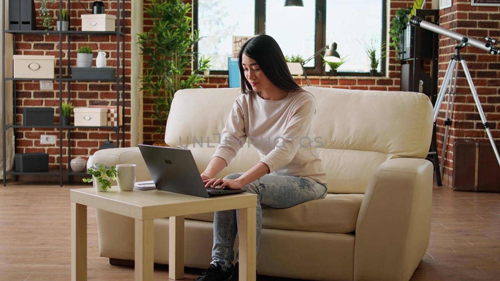 Attractive asian freelancer doing remote work on modern portable computer. Focused smiling heartily young adult person sitting on sofa at home while working remotely on laptop.
