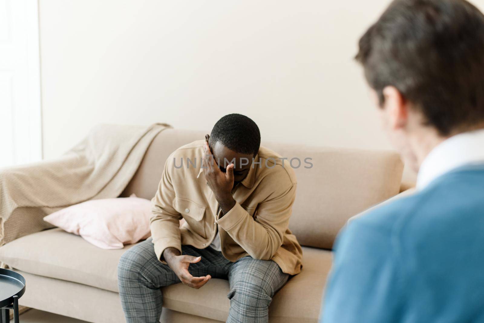 Frustrated young african american man sitting on sofa and holding hands near face in office at session with psychologist or psychiatrist. Desperate black man suffering from nervous breakdown or depression, having session at psychologist.