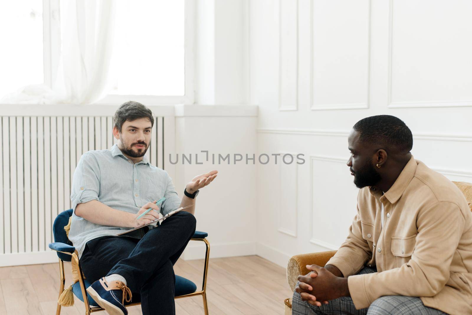 A male psychologist conducts psychological therapy with an African American man, makes notes in a clipboard