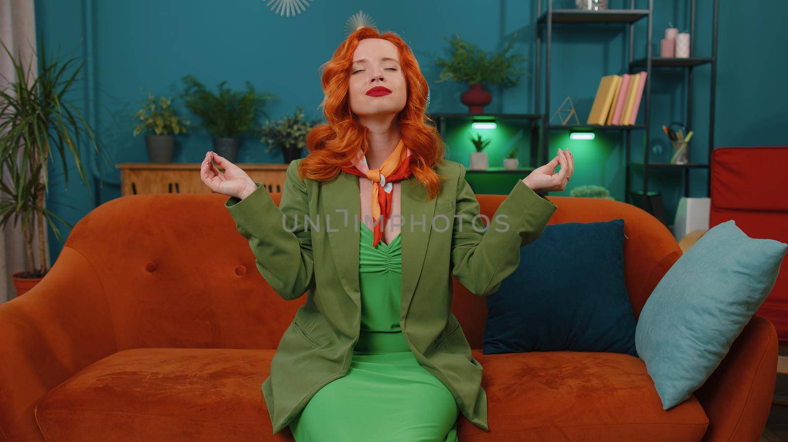 Keep calm down, relax, inner balance. Woman breathes deeply with mudra gesture, eyes closed meditating with concentrated thoughts, peaceful mind. Redhead adult girl sit at home in living room on couch