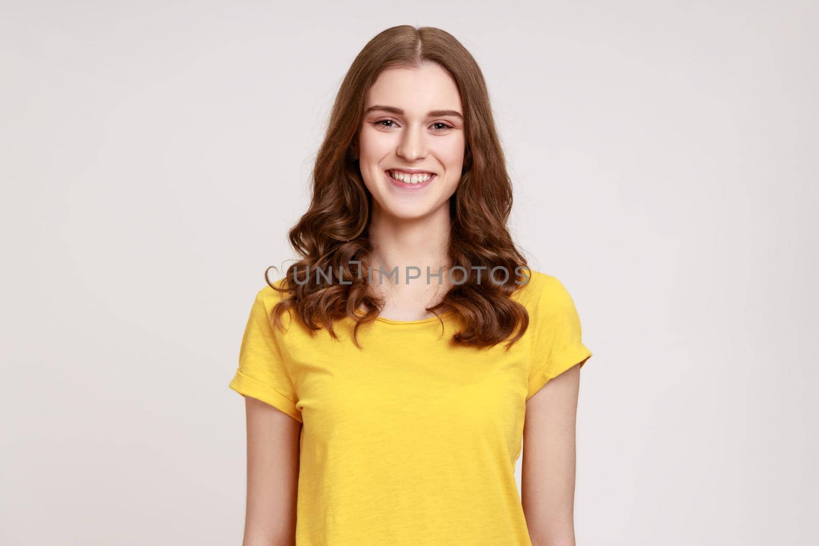 Portrait of cheerful teenager girl in yellow casual T-shirt sincerely smiling at camera, positive mood, being glad to meet somebody. Indoor studio shot isolated on gray background.
