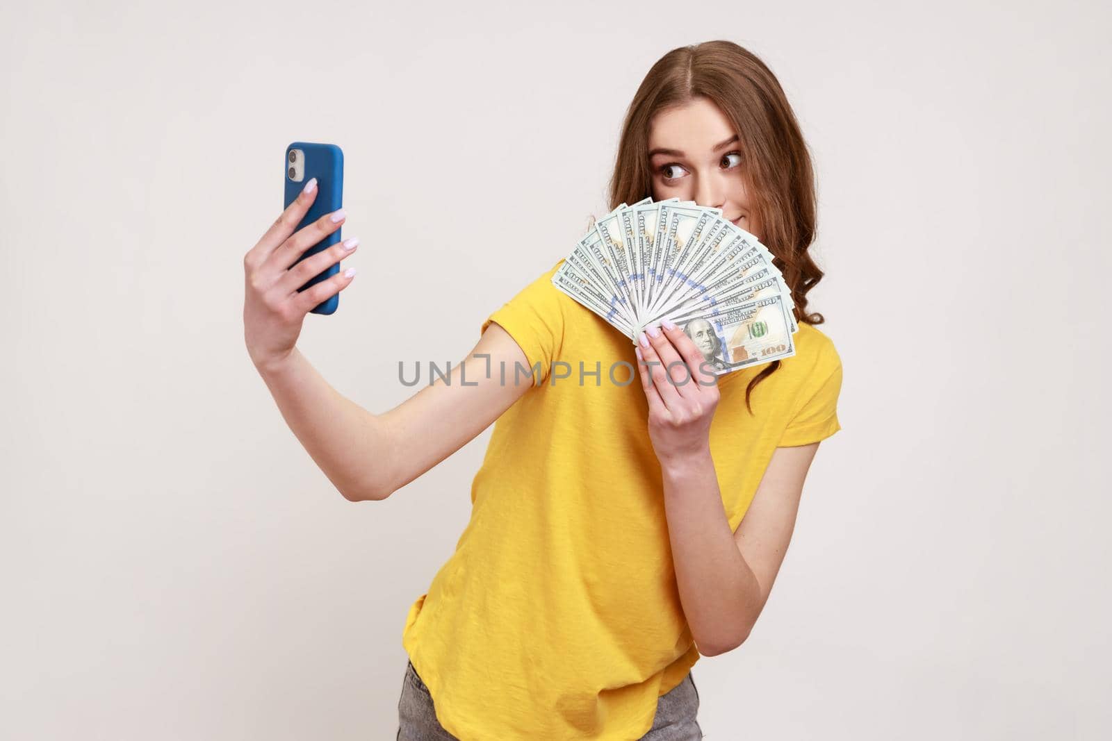 Portrait of rich teen girl with brown hair in T-shirt holding money and taking selfie by mobile phone, boasting of big profit, successful online betting. Indoor studio shot isolated on gray background