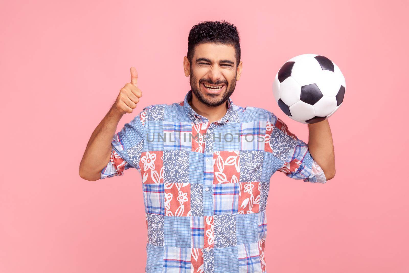 Happy excited bearded man with dark hair and beard wearing blue shirt posing with soccer ball and showing thumb up, cheering on football match. Indoor studio shot isolated on pink background.