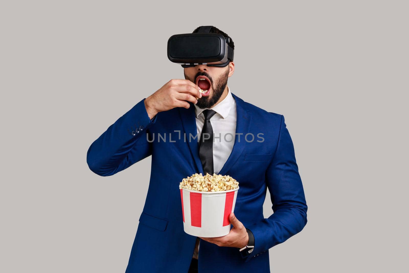 Portrait of excited amazed bearded businessman in VR headset watching movie with popcorn, eating tasty snack, wearing official style suit. Indoor studio shot isolated on gray background.