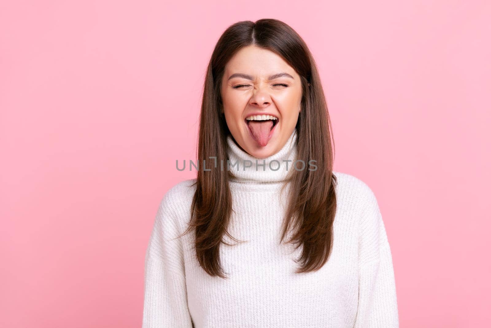 Childish woman with dark hair showing tongue out, expressing positive emotions, having playful mood, wearing white casual style sweater. Indoor studio shot isolated on pink background.